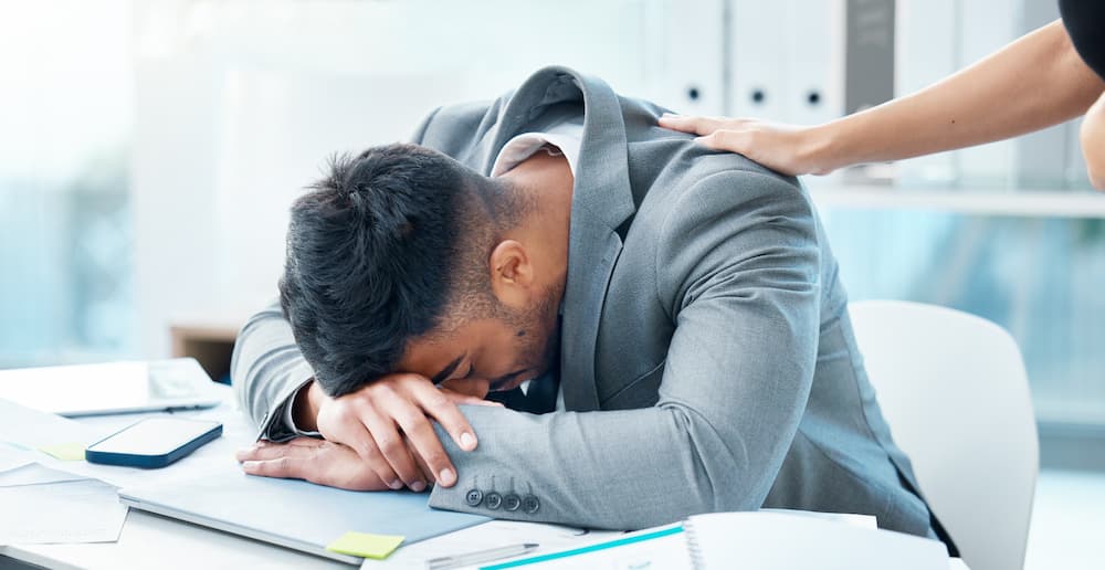 A man in a grey suit with head down in his hands because of career challenges.
